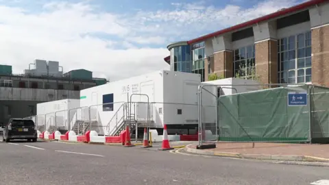 White mobile buildings in a lay-by or parking area on a hospital campus, with the main hospital buildings surrounding them
