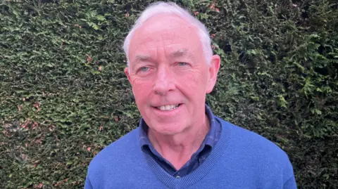 Pete Russ smiles at the camera, standing in front of a hedge. He wears a blue jumper and shirt.