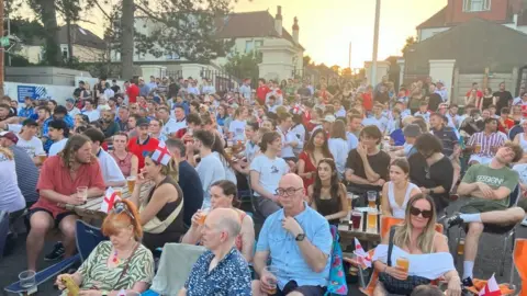 Hundreds of people are sitting in a fanzone in the car park at the Memorial Stadium in Bristol. Many of them are wearing England shirts or holding small England flags
