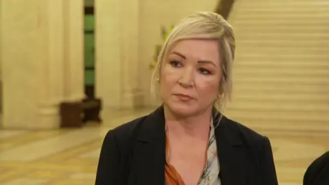 O'Neill has blonde hair tied back. She is wearing a black blazer with a patterned blouse underneath.