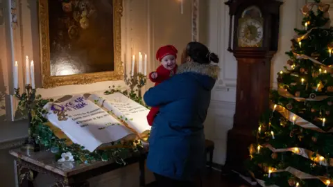 National Trust One of the rooms in Mompesson House with a mother holding a baby and looking at an oversized book of fairytales laid out on a table with candles and a Christmas tree and decorations in the background