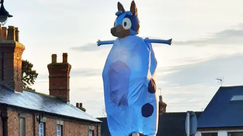 A statue covered in a "fancy dress" costume as Bluey the cartoon dog. A sheet-like costume covers the stone statue, with a modelled head depicting the character on top. Two arms stick out at right angles. Surrounding rooftops are also visible. 