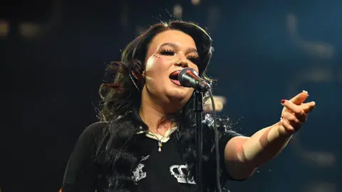 Getty Images Lola Young performing at the Spotify Best New Artist show, she is wearing a black short sleeved t-shirt. She has long wavy black hair and is singing into a mic. 