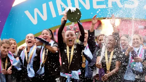 PA Media Manchester City's Alex Greenwood lifts the trophy. She wears a black sports top. Other players behind her appear to shout in celebration, and some are spraying liquid from bottles.