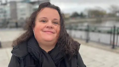 A woman with long dark hair wearing a black scarf with white specks and a black coat.