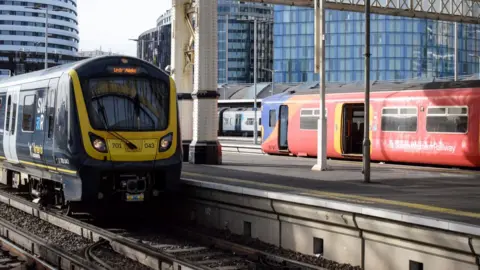 A red and yellow train with South West Railway branding at a station with another behind it