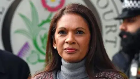 Gina Miller, who has long dark hair and is wearing a grey polo-neck jumper under a collarless jacket. Behind her can be glimpsed a bearded police officer. The logo for the UK Supreme Court is directly behind her. 