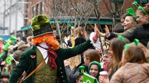 Pacemaker A man wears a green, velvet leprechaun outfit, complete with a large, ginger beard and a pipe high-fives children who are lining up along the street in Belfast. 