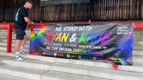 A man in a black t-shirt stands on concrete steps to the left of a banner secured to a railing. The banner has a rainbow gradient design and bold text which reads: WE STAND WITH RYAN & KAUE.