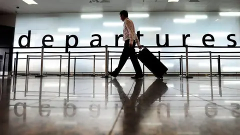 A man walking along through departures at Glasgow airport, carrying a wheeled trolley bag.