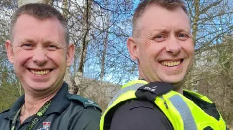 Scottish Ambulance Service Two twin brother with short cropped hair, one is in a Scottish Ambulance Service T-Shirt, the other is in Police Scotland top and high visibility vest