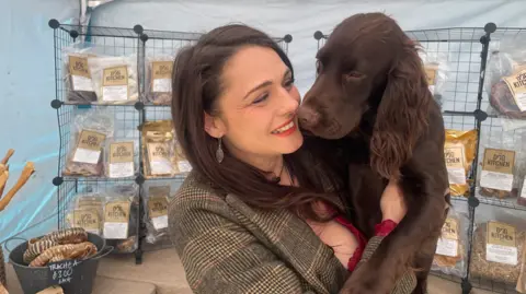 A woman with long brown hair and a tweed jacket holding a brown dog with racks holding plastic packages behind her