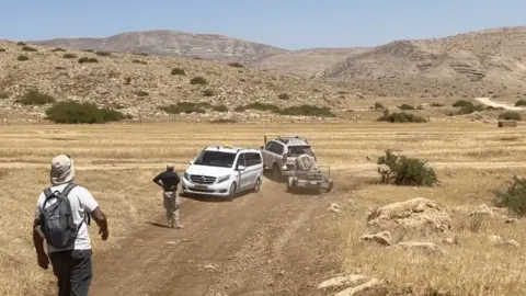 A white jeep blocks a white people carrier-type vehicle on a dirt road