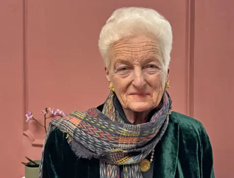 A woman with short grey hair and gold earrings - wearing a patterned scarf - looks straight at the camera