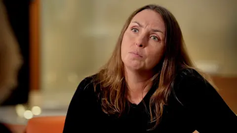 Emma Wilson frowns slightly at the camera. She has long, slightly wavy, brown hair and is wearing a black v-neck top. She is sitting on some orange furniture and is against a blurred orange and brown background.