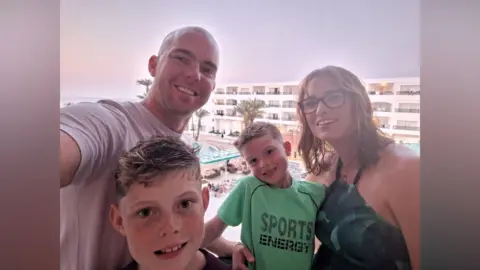 Family handout A selfie of a family from a balcony at a hotel. The sky is a grey/purple colour. There is a man, woman and two young boys. 