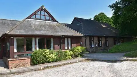 Dave Percival A small redbrick building with a triangular roof and large windows, next to a road and a grass verge. It's a sunny day and the sky is blue