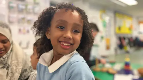 A girl with curly hair is smiling at the camera in a light blue jumper 