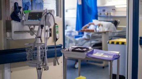 Medical equipment stands in front of a hospital room with a patient out of focus inside the room
