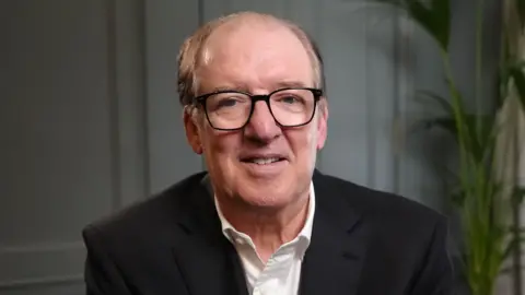 Sir David Murray sits in front of a pale blue wall wearing a black blazer and a white shirt. He has black rimmed glasses and is balding, with sandy hair at the sides. He is smiling slightly.