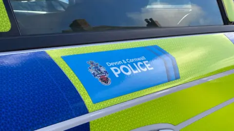 A picture of the side of a green and blue police car. The blue Devon and Cornwall Police logo is pictured on the side of the vehicle. 