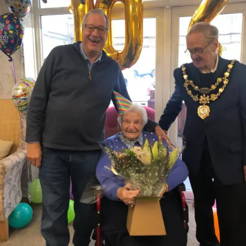 Two men and a woman at a birthday party, with balloons in the background. The man on the left has grey hair and glasses, and he is wearing a blue jumper and jeans. The man on the right has grey hair and glasses, and he is wearing large gold ceremonial necklaces over a dark jacket. The woman, who is sitting down, has grey hair and glasses. She is wearing a stripey cone-shaped party hat and a purple jumper.