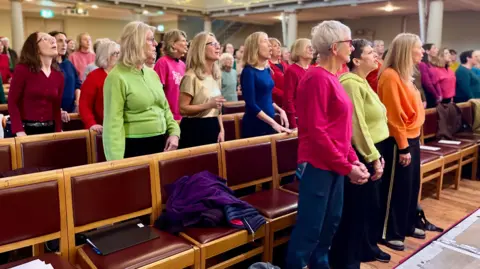 A group of people wearing colourful tops singing together but from the front left angle