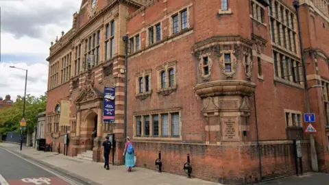 A large red brick building with small, black framed windows and people walking outside