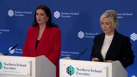 Michelle ONeill - who has shoulder-length blonde hair- in a black suit and Emma Little Pengelly - who has longer dark brown hair - in a red suit , standing at a press podium. The background is blue and has NI executive logos across it.