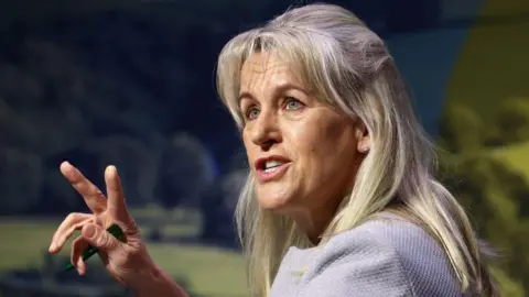 Getty Images A close-up head-and-shoulders shot of Baroness Batters as she raise two fingers to illustrate a point while making a speech. The background is a soft-focus shot of the countryside