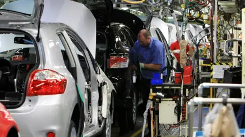PA Media A man in a blue shirt working on a production line in a car factory. There are several part assembled cars in a line with machinery around them.