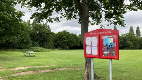 Shariqua Ahmed/BBC A red 'Bretton community' noticeboard with green grass and trees in the background.