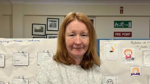 Helen Smyth está de pie, con el pelo rojizo hasta los hombros. Lleva un jersey de punto color crema. Detrás de ella hay una cartulina con dibujos de ella a mano en un lado y texto escrito en el otro. A lo lejos, otra pared con imágenes en blanco y negro y una señal de salida de incendios.