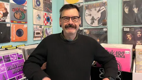Gary Matfin standing in front of a display of vinyl records on the wall of his shop. He has dark brown hair and is wearing glasses and a black jumper.