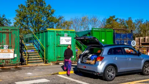 A lady is standing at the boot of the car and looking towards four storage containers that are used for waste at a recycling centre. 