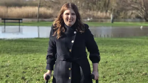 A woman with mid-length brown hair, standing outside on grass in a black jacket. She is smiling widely and her hands clasp crutches. Behind her is a blurred patch of water and some trees.