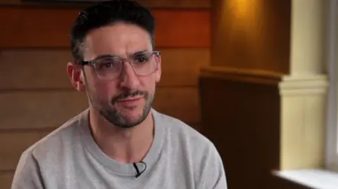 BBC Yoni Finlay stares forward while wearing a grey sweatshirt. He has black hair and is wearing transparent glasses. 