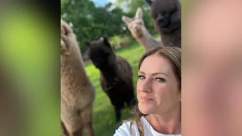 Tamsin Harrison Tamsin Harrison is smiling to the camera with four alpaca's behind her on a field. Two alpacas are black and fluffy and the other two are white. 