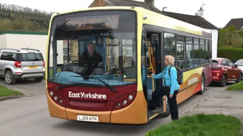 A women with short blonde hair, wearing a light blue sweater, black trousers and a black rucksack getting on an East Yorkshire bus called the Wolds Explorer. The bus has orange, burgundy and yellow markings and is parked on the side of a residential street. 