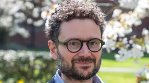 University of Cambridge Prof Paolo Campana is looking at the camera. He has curly dark hair and facial hair. He is wearing dark-rimmed spectacles and a blue shirt and jacket. He is standing in a garden, which is blurred in the background.