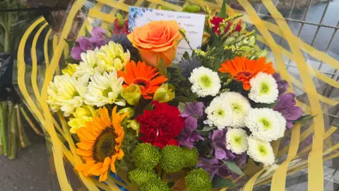 A close up of a bouquet of flowers which are orange, white, green, red and purple. The blue and white note card is poking up from behind the heads of the flowers. The top line reads: "Rest in peace." It is wrapped in clear plastic with a yellow striped design on it.