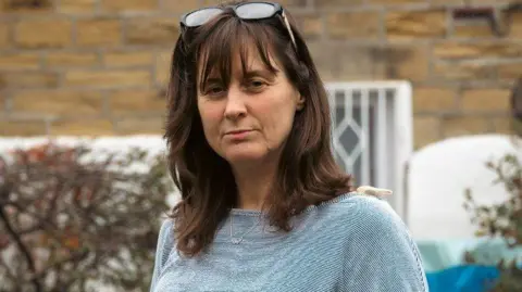 A woman with shoulder length brown hair. She has glasses resting on her head and is staring straight ahead. She is wearing a light blue top and is stood in front of a bricked building. 