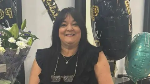 Louisa Curtis is wearing a sleeveless black top with ruffled detailing and has a pair of glasses hanging from a chain around the neck. Jewellery including a necklace, bracelet, ring and wristwatch, is visible. 