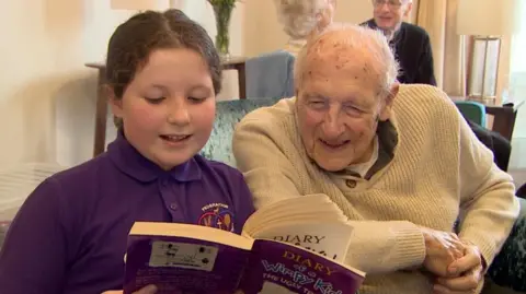 Jim Earle sits beside Eliza as she reads from a book. He is looking at the page that she is reading from. He is wearing a white jumper and she is wearing a purple polo shirt. They are sitting in a living room. 