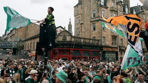Dozens of Celtic fans, many wearing replica kits or scarves and waving club flags, gather in a city centre area.
