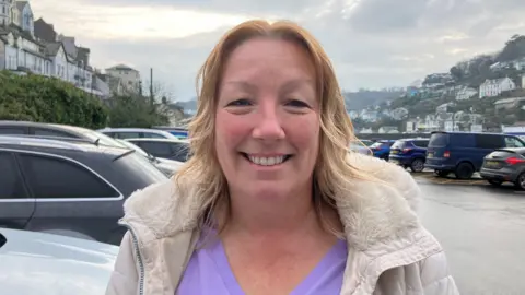 Woman with mid-length blonde hair smiling at the camera. She is in a car park in Looe and is wearing a fur lined hooded coat and a purple top