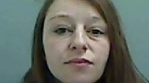 A grainy head and shoulders image of Natalie Jenkins who has long brown hair. She is looking directly down the camera lens.