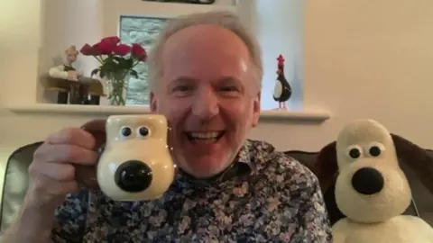 Nick Park has grey hair and is wearing a floral shirt. He is sat on a sofa with a Gromit sift toy next to him and is holding a Gromit shaped mug. On a window ledge behind is a vase of red flowers, a penguin character, and the Wallace character from his animations.