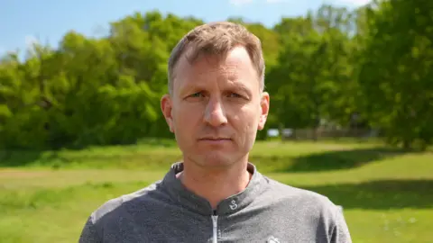 Qays Najm/BBC A head and shoulders of a man. He is wearing a grey top, standing in an open green space with trees behind him, and looking directly into the camera.