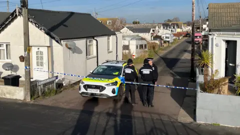 Shaun Whitmore/BBC Police officers guard a cordon on a narrow road in Jaywick. There is a marked police car next to them and blue and white tape. There are several small properties and caravans lining the road.